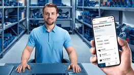 A man in a blue shirt sits at a table and looks into the camera. In one hand, he holds a smartphone displaying an app. In the background, there are shelves with Bosch Professional tools. The scene conveys a professional workshop atmosphere.