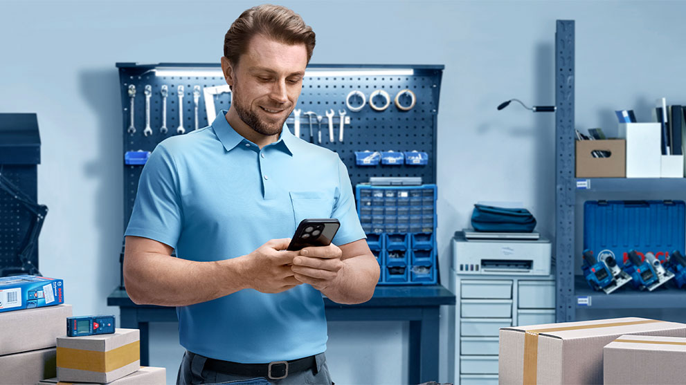 A man in a blue shirt stands at a table, looking at his smartphone. Around him are boxes, a toolbox, and a workbench in the background.