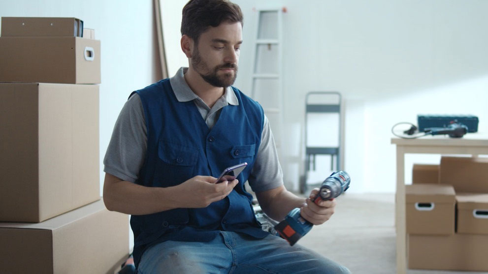 A man in a blue vest is sitting on the floor holding a power drill and a smartphone. In the background, there are boxes and a ladder.