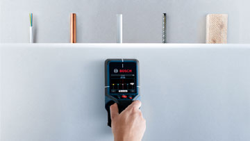A hand holds a Bosch Professional measuring device in front of a wall with pens.