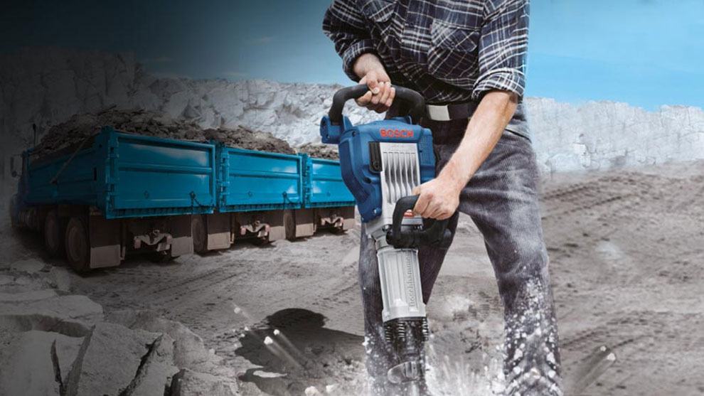 A man is using a blue Bosch Professional demolition hammer in gravel.