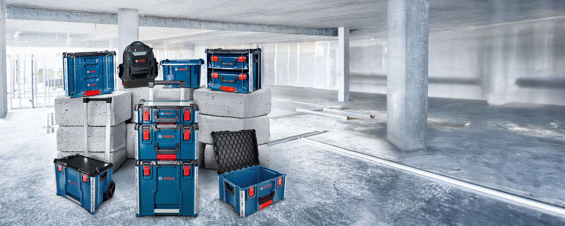 A workspace with several blue toolboxes from Bosch, arranged on gray concrete blocks. The floor is made of smooth gray concrete, and there are some construction materials in the background. A black bag is also visible. The mood is industrial and functional.