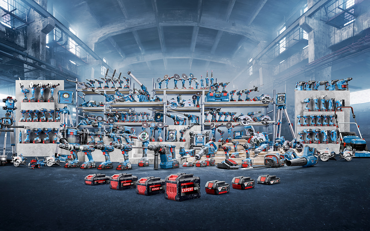 A variety of blue Bosch Professional tools is arranged on shelves and the floor in an industrial hall. Several battery tools, machines, and accessories are visible, including drills, grinders, and batteries labeled "EXPERT". The background is foggy and provides an industrial ambiance.