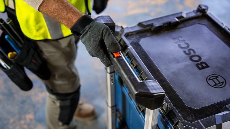 A worker in a safety vest grabs the handle of a blue Bosch toolbox on a cart.